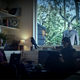 Two players of The Past Within in their living room, while a deer skull-headed man looks in the window.